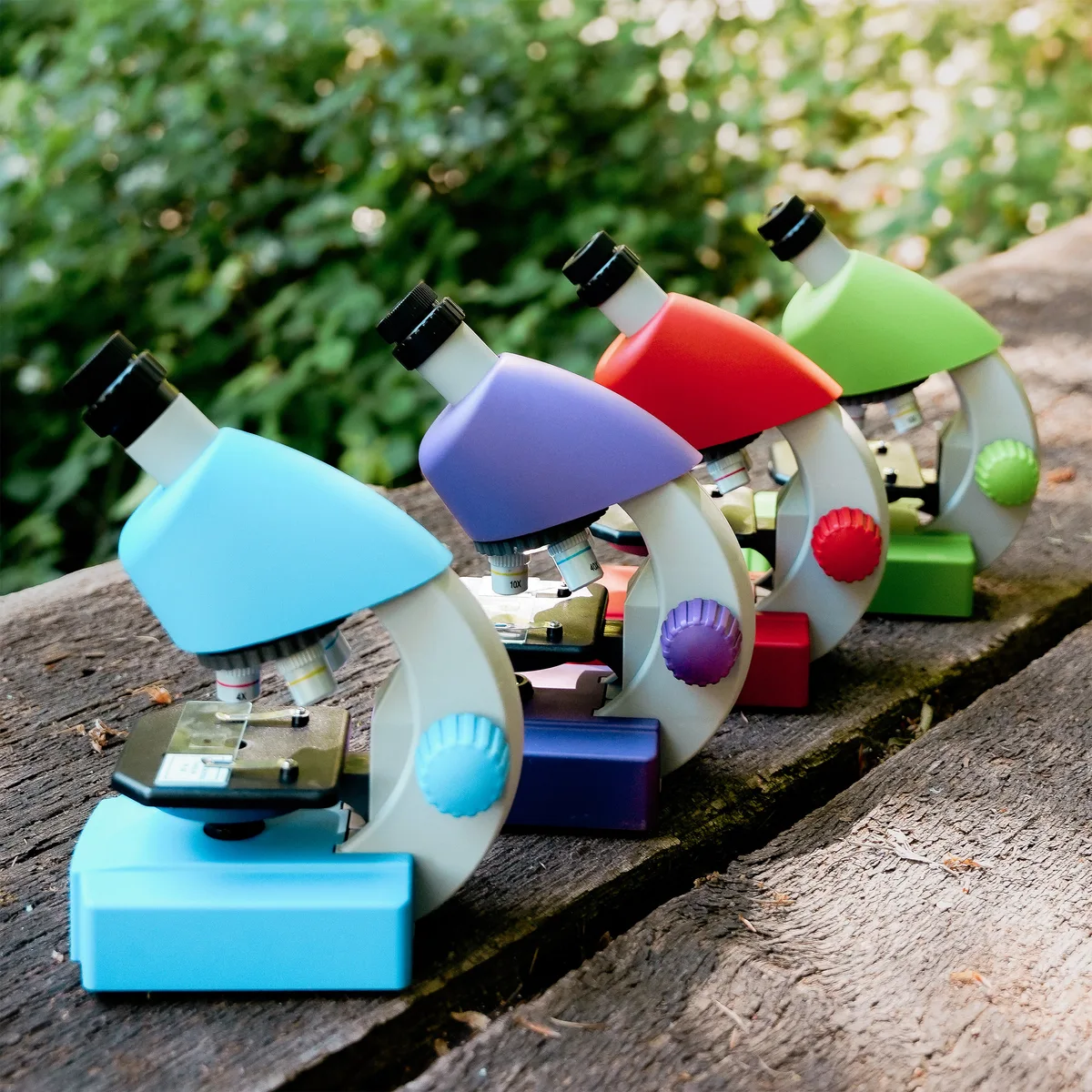 Quatre microscopes Bresser Junior de couleurs différentes posés sur un banc en bois